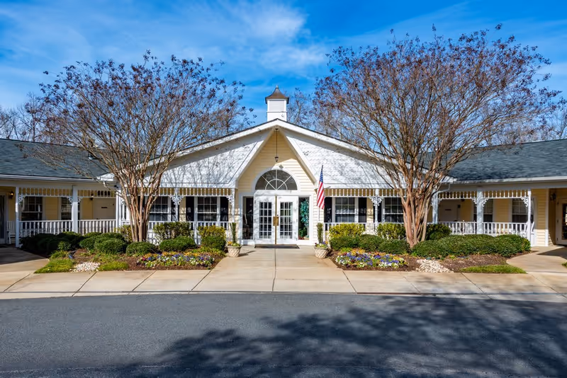 Front entrance of a single-story senior living building with symmetrical porches, landscaping, and an American flag under a blue sky.
