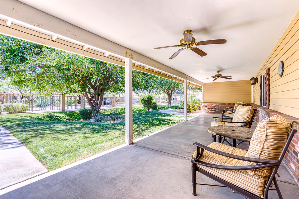 Covered porch with ceiling fans and cushioned chairs overlooking a grassy courtyard with trees and a fence.