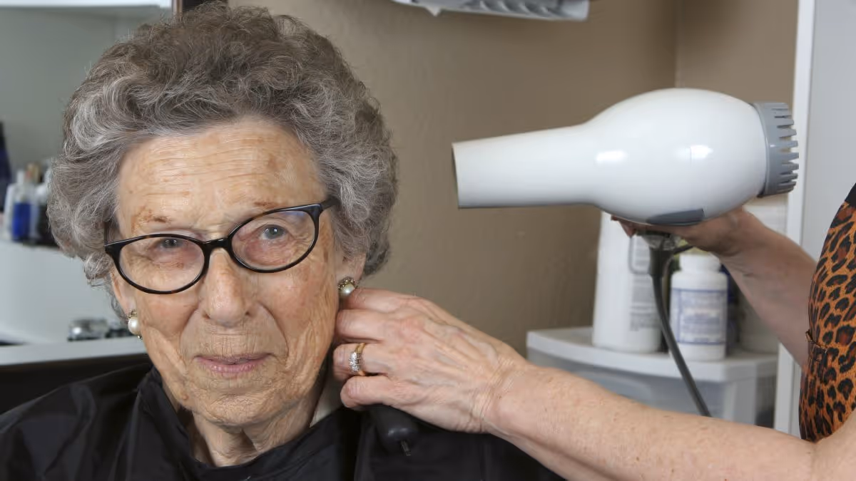 An elderly woman with glasses and pearl earrings is having her hair dried with a white hairdryer by another person wearing a leopard print top in a salon-like setting.
