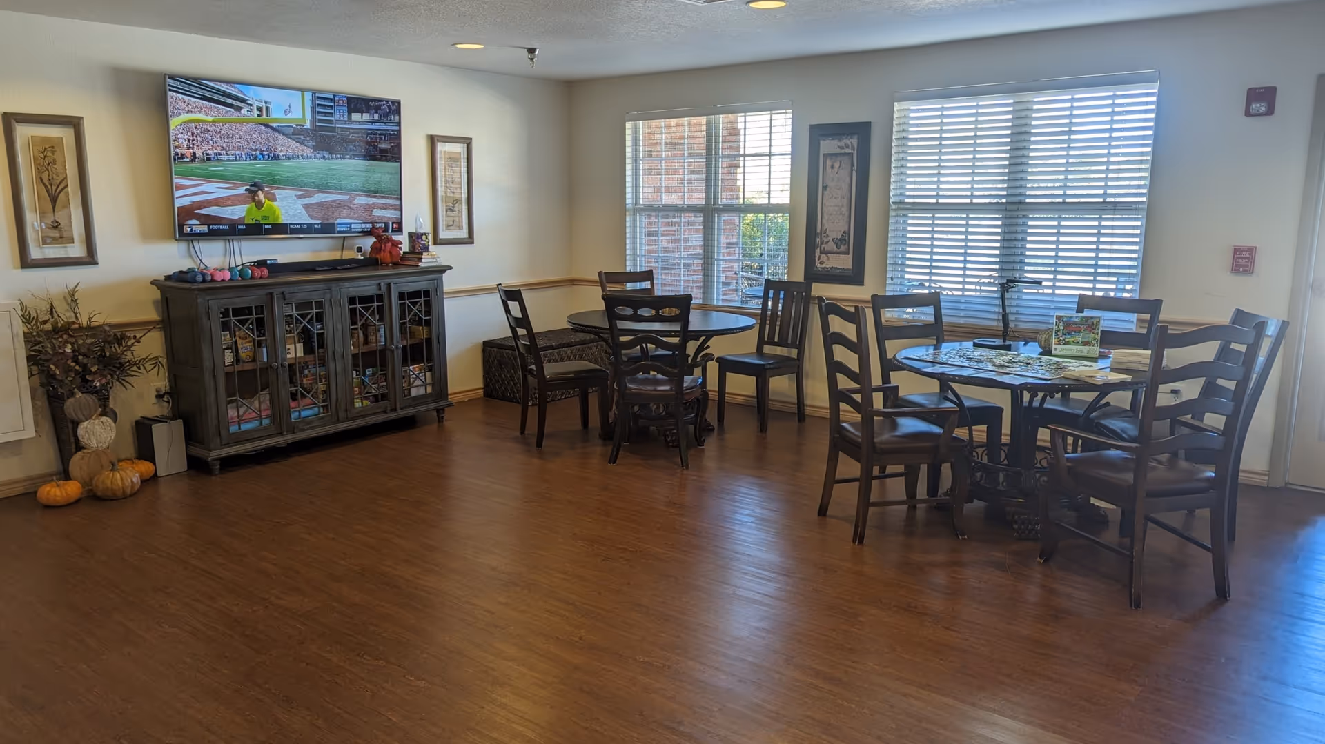 Spacious common room with wooden floors, round dining tables and chairs, a wall-mounted TV and a sideboard near large windows.