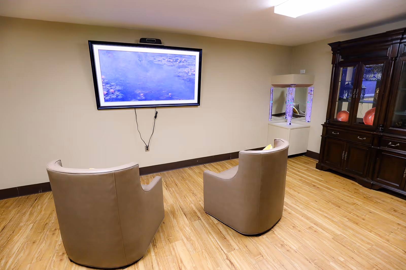 A small room with two modern beige armchairs facing a wall-mounted flat screen TV displaying a blue water-themed image. The room has light wooden flooring, beige walls, a dark wooden cabinet with glass doors on the right, and a small illuminated bubble aquarium next to the cabinet.
