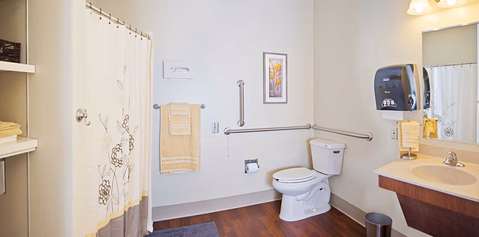 A clean and accessible bathroom featuring a toilet with grab bars on the walls, a shower with a floral-patterned curtain, a sink with a mirror above it, a paper towel dispenser, and neatly folded towels on a shelf.