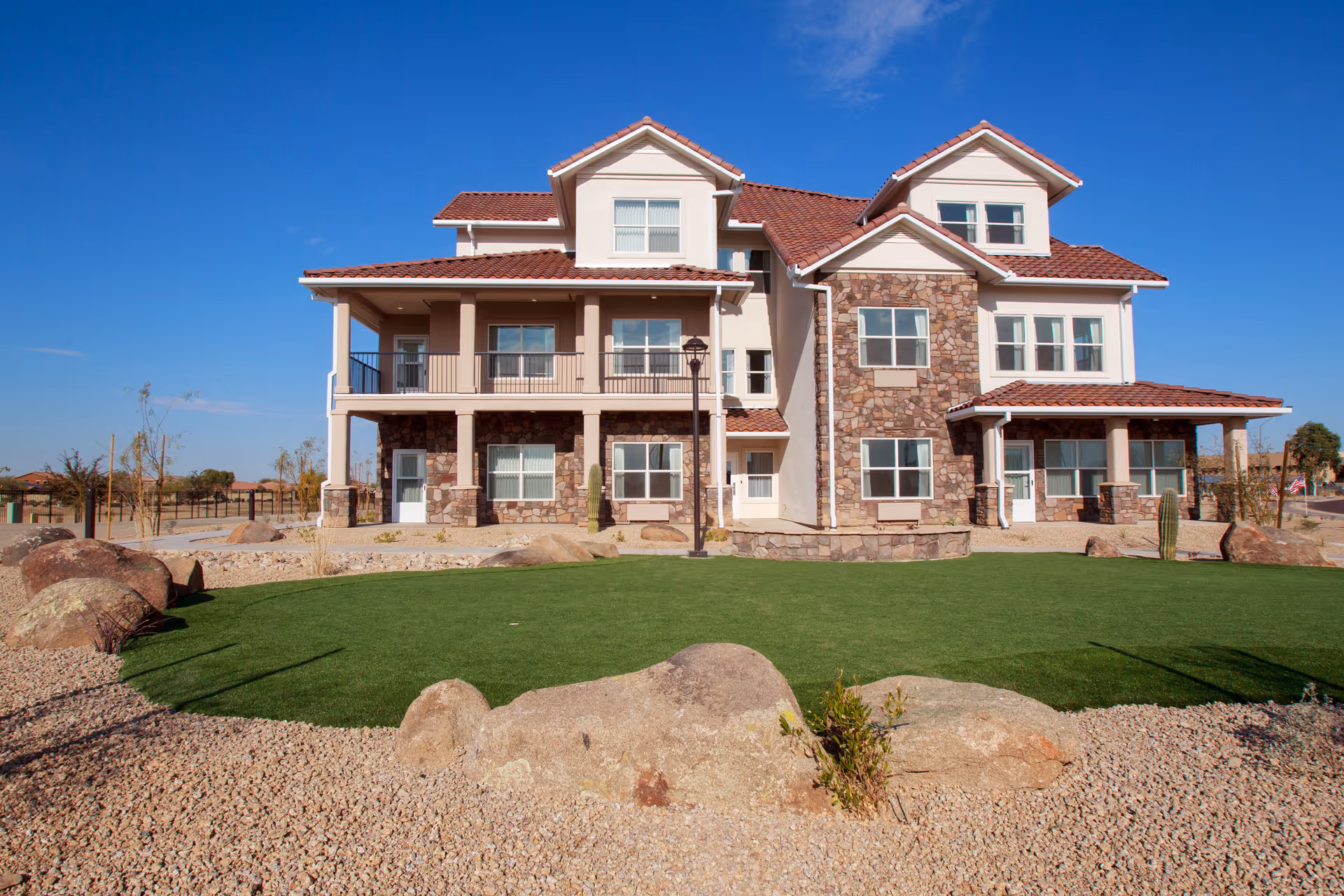 Exterior view of a large, multi-story residential building with stone and stucco facade, red tile roof, balconies, and a well-maintained lawn with rocks and desert landscaping under a clear blue sky.