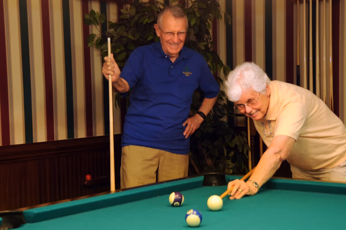 An elderly man and woman playing pool indoors. The man is standing and smiling while holding a pool cue, and the woman is leaning over the pool table aiming to hit the cue ball. The room has striped wallpaper and a plant in the background.