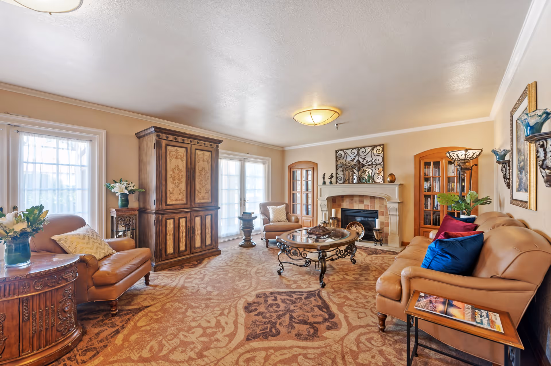 A cozy living room in Katella Senior Living Community featuring a tan leather sofa with colorful pillows, a matching armchair, a glass-top coffee table with ornate metal legs, a fireplace with decorative items on the mantel, wooden cabinets with glass doors, and large windows with sheer curtains allowing natural light to fill the room. The room has warm beige walls and a patterned carpet.