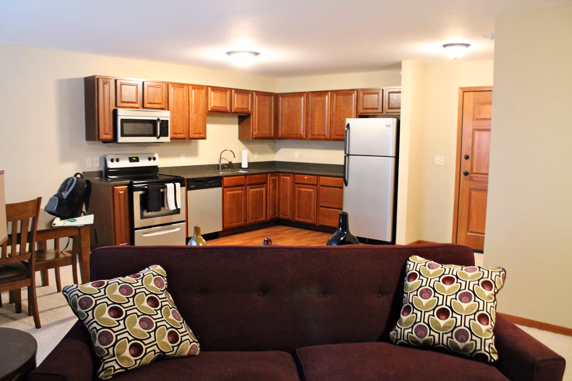 Open-plan living area with a purple sofa and patterned pillows facing a kitchen with wooden cabinets and stainless steel appliances.