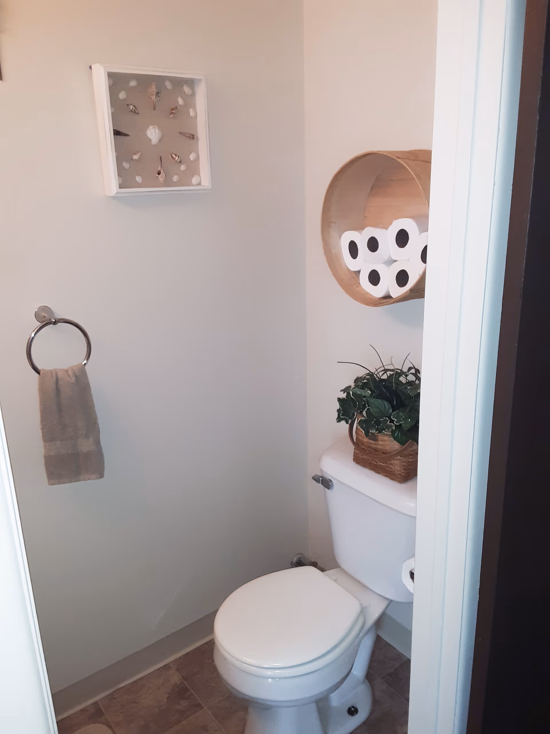A small bathroom with a white toilet, a wicker basket with green plants on top of the toilet tank, a round wooden shelf mounted on the wall holding six rolls of toilet paper, a towel ring with a beige towel, and a framed decorative piece with seashells on the wall.