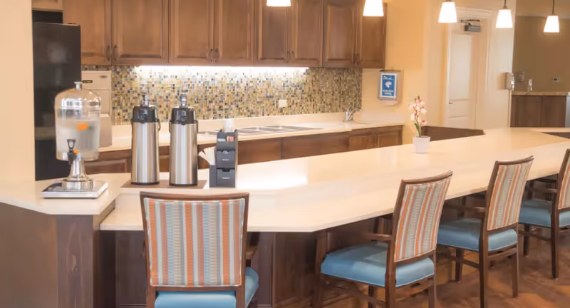 A modern kitchen area with a long white countertop and wooden cabinets. There are several chairs with striped backs and blue seats lined up along the counter. On the counter, there are two large beverage dispensers, a small container with stirrers or straws, and a small potted plant. The backsplash features a mosaic tile design, and pendant lights hang from the ceiling above the counter.