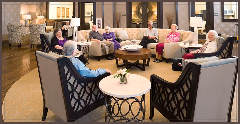 A group of elderly residents seated in a circle on sofas and armchairs in a bright communal living room.