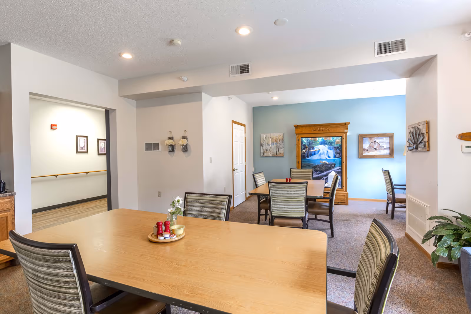 Communal dining room with several wooden tables and striped upholstered chairs, a light blue accent wall with framed artwork, and a hallway entrance.