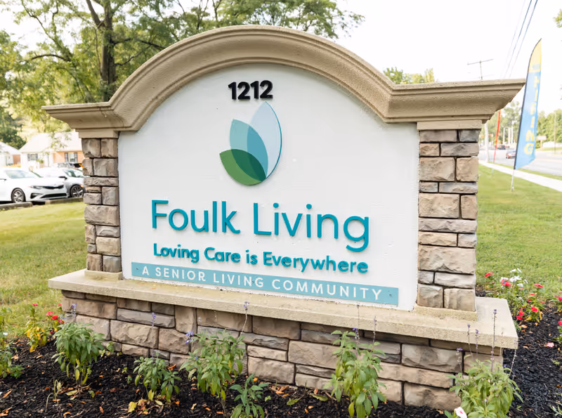Outdoor stone and concrete sign for Foulk Living senior living community with the address number 1212, a green and blue leaf logo, and the slogan 'Loving Care is Everywhere'. The sign is surrounded by small plants and flowers with a street and cars visible in the background.