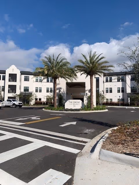 Exterior view of The Grove at Trelago senior living facility showing a three-story white building with multiple windows, two tall palm trees in front of the entrance, a sign with the facility name, and a parking area with marked lanes and a white vehicle parked on the left side under a partly cloudy blue sky.
