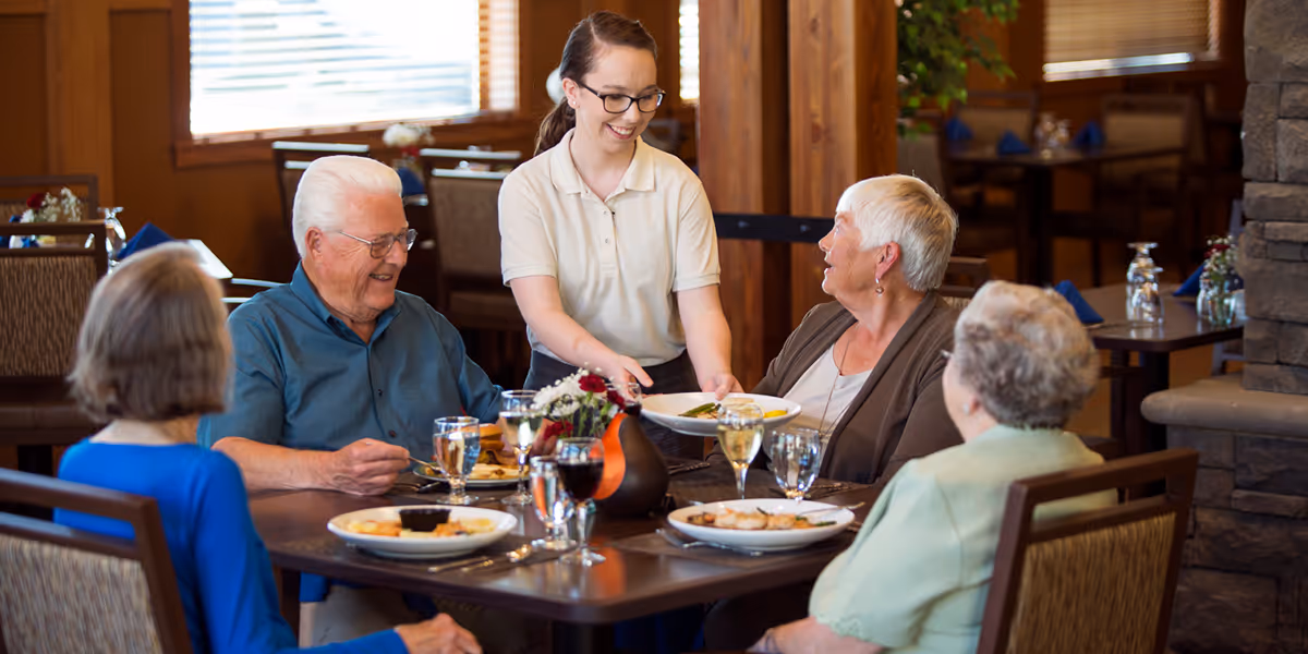 A young female server smiling and serving food to a group of four elderly people seated around a dining table in a warm, wood-accented dining room at Dallas Retirement Village Assisted Living.