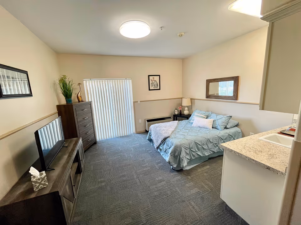 A cozy bedroom in an assisted living facility featuring a bed with blue bedding and pillows, a nightstand with a lamp and decorative items, a dresser with a plant and decorative sculpture, a flat-screen TV on a wooden stand, a kitchenette area with a sink and countertop, and vertical blinds covering a sliding glass door.
