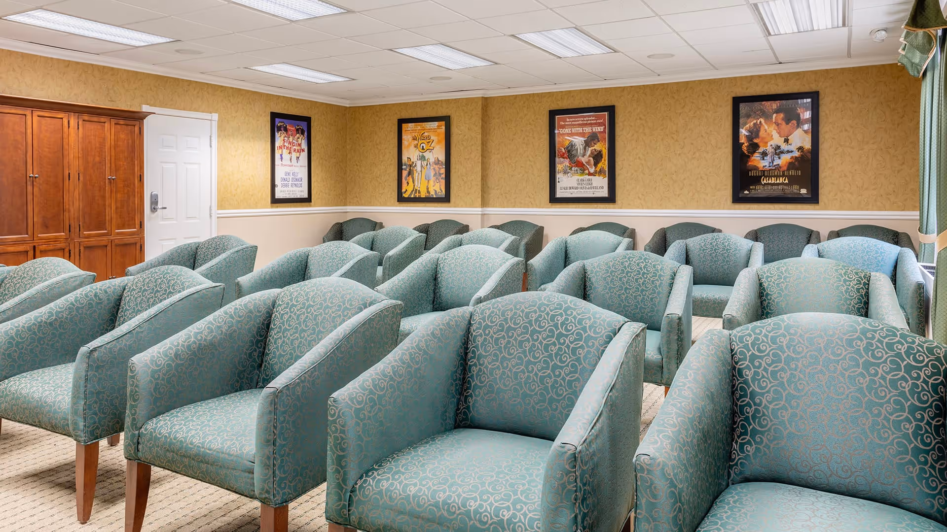 A room with multiple rows of green patterned armchairs arranged facing forward, resembling a small theater or presentation room. The walls are decorated with framed movie posters, and there is a wooden cabinet and a white door on the left side.