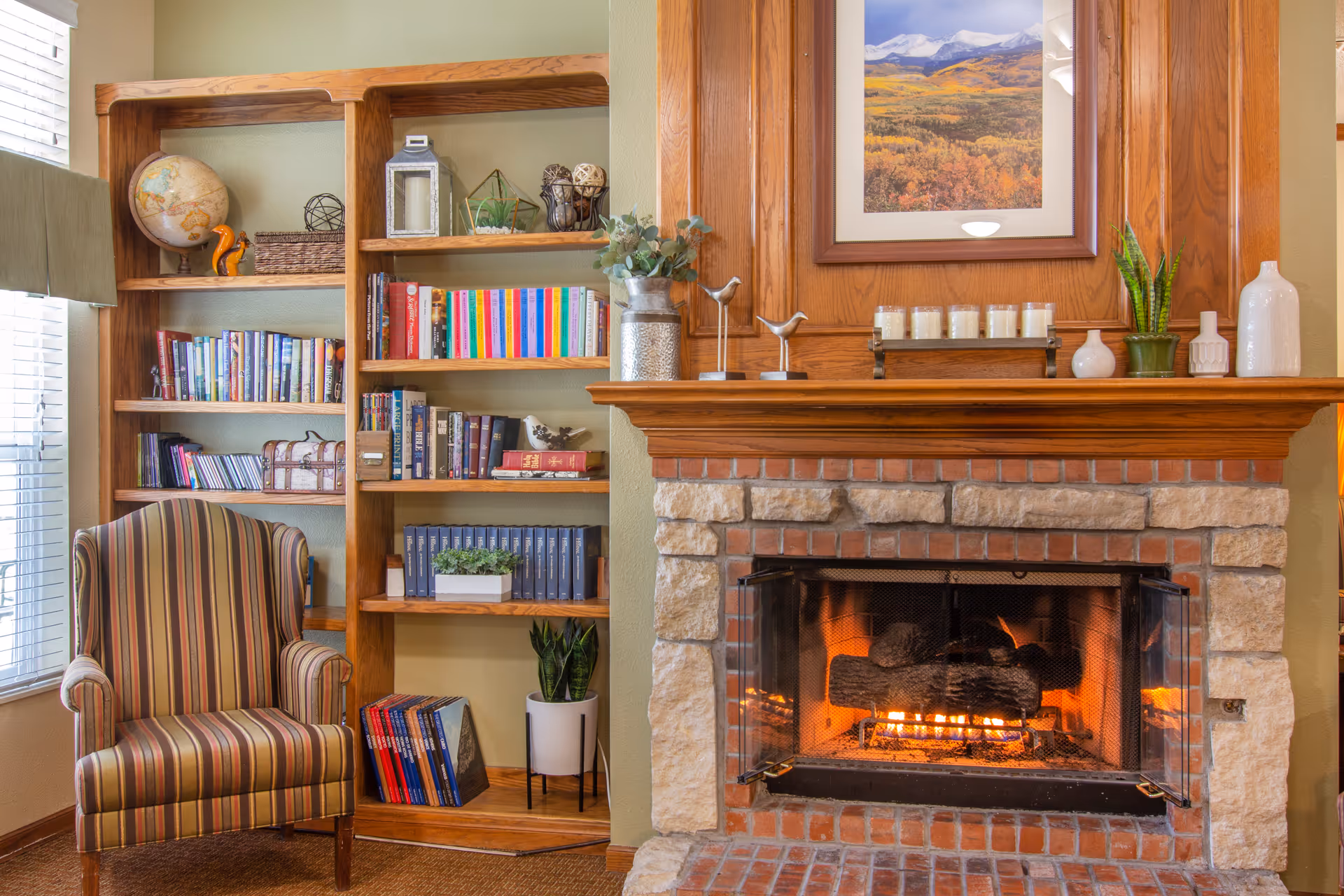 Cozy living room featuring a striped armchair, wooden bookshelf, and a lit stone fireplace beneath a framed landscape.