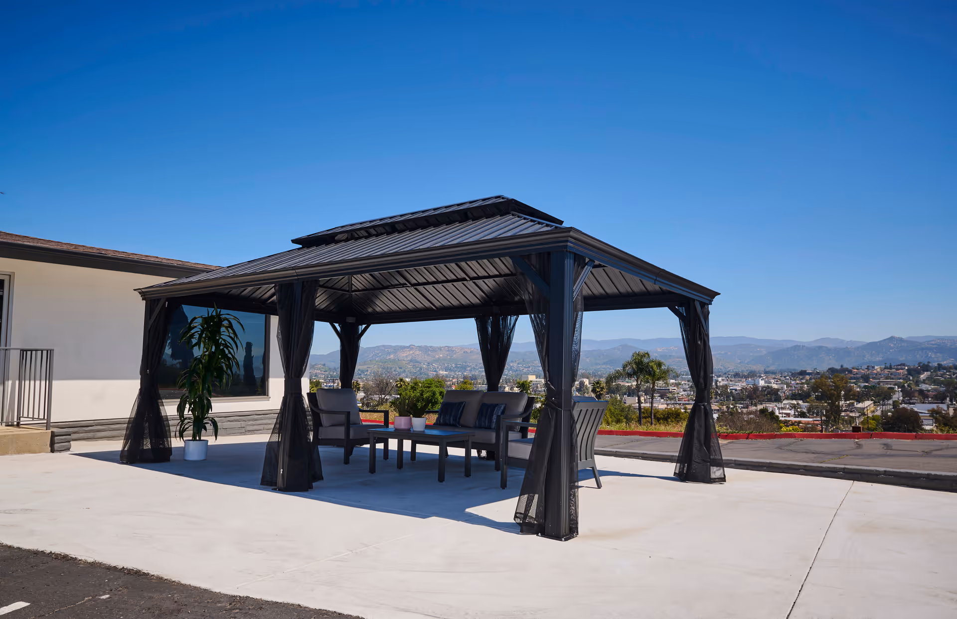 Outdoor covered gazebo with seating on a concrete patio overlooking a city and distant hills.