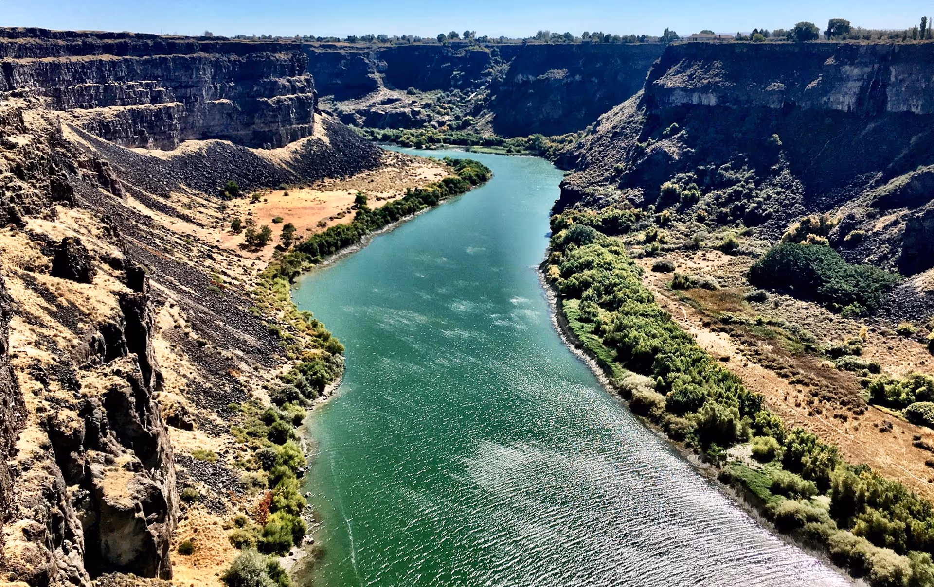 A wide river flowing through a deep canyon with steep rocky cliffs on both sides. The landscape is dry with sparse vegetation on the cliffs and greener bushes along the riverbanks under a clear blue sky.