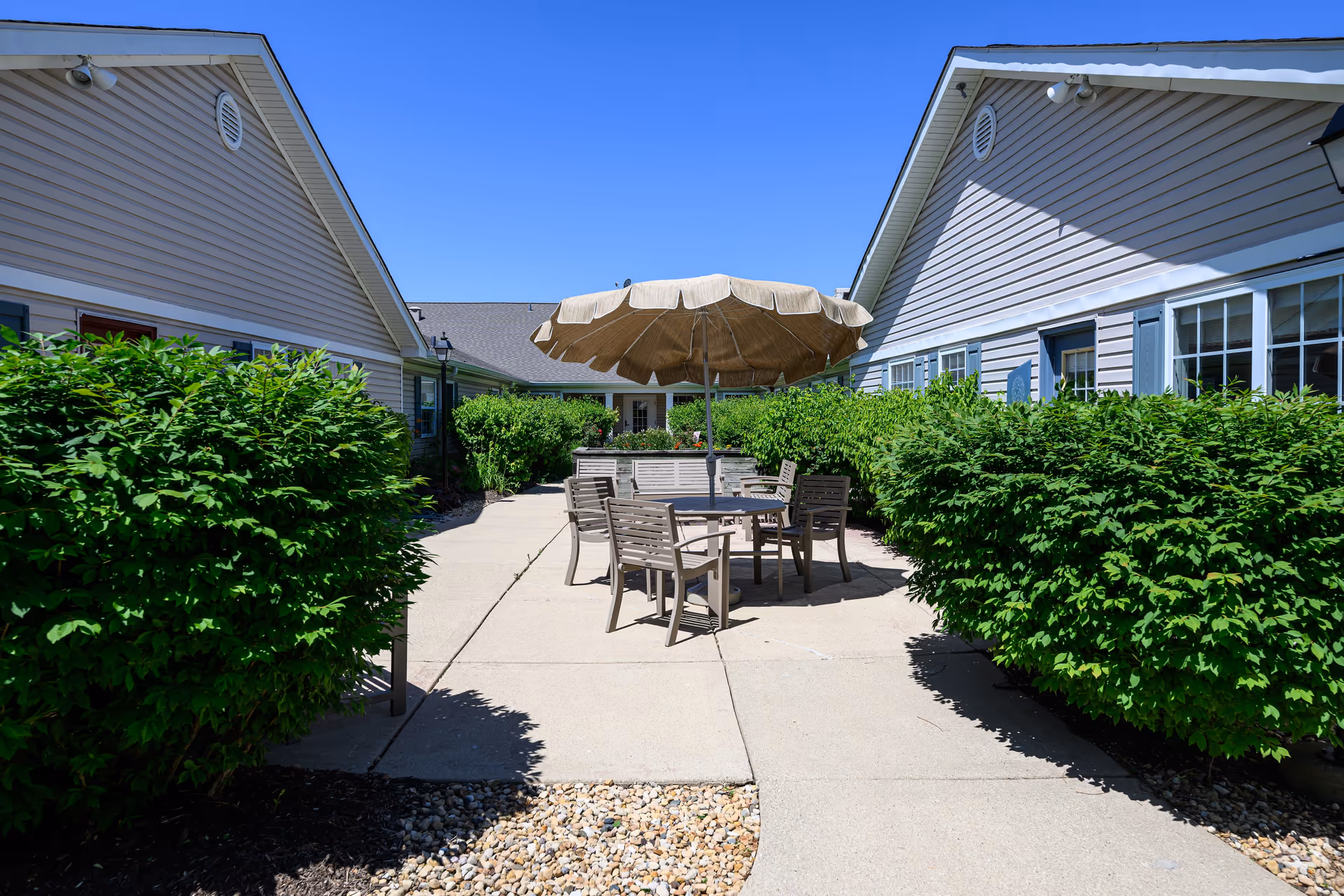 Outdoor patio area between two buildings with a round table, several chairs, and a large beige umbrella. The patio is surrounded by green bushes and the sky is clear and blue.