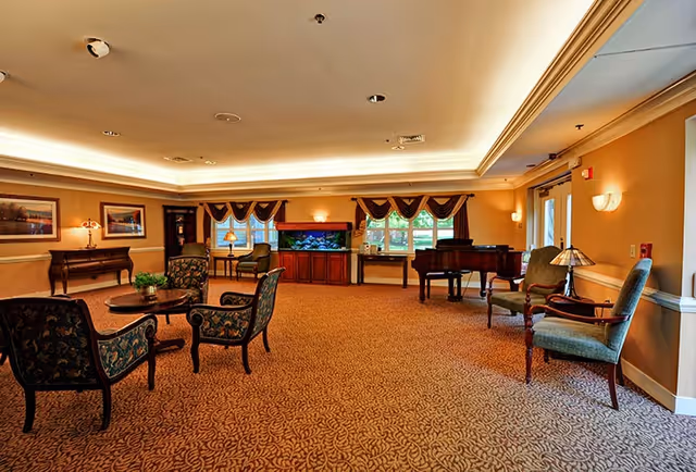 Spacious senior living common room with grouped seating, a piano, an aquarium, and warm ambient lighting.
