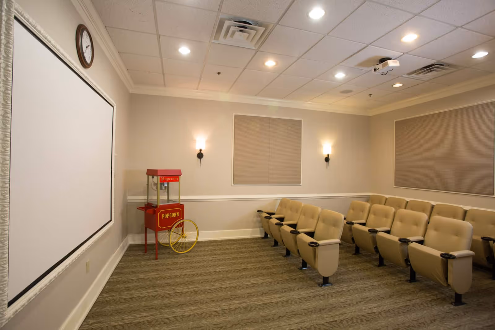 A small theater room with beige cushioned seats arranged in rows facing a large white projection screen. There is a red popcorn machine with yellow wheels in the corner of the room. The walls are light-colored with two wall sconces and two covered windows. A clock is mounted above the screen, and a ceiling-mounted projector is visible.