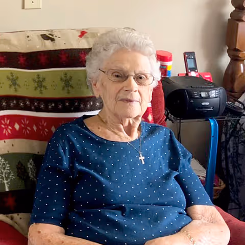 An elderly woman with short white hair and glasses sitting on a chair covered with a festive blanket featuring Christmas tree and snowflake patterns. She is wearing a blue shirt with small white polka dots and a cross necklace. Behind her, there is a small table with a black radio, a red container, and a cordless phone.