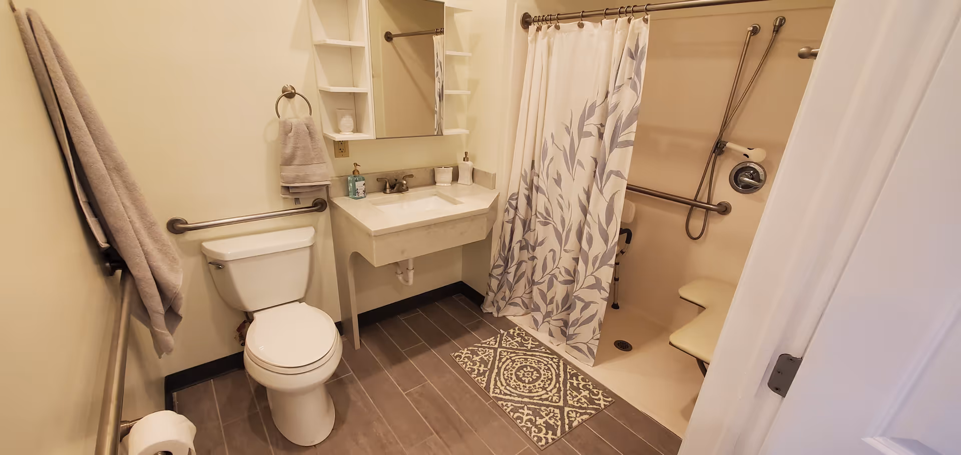 Accessible bathroom with a toilet, wall-mounted sink, grab bars, and a shower with curtain and fold-down seat.