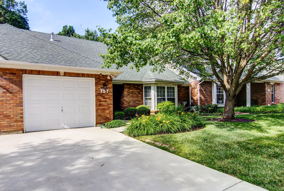 Exterior view of a brick residential building with a white garage door numbered 757, a concrete driveway, green lawn, flowering plants, and a large tree providing shade.