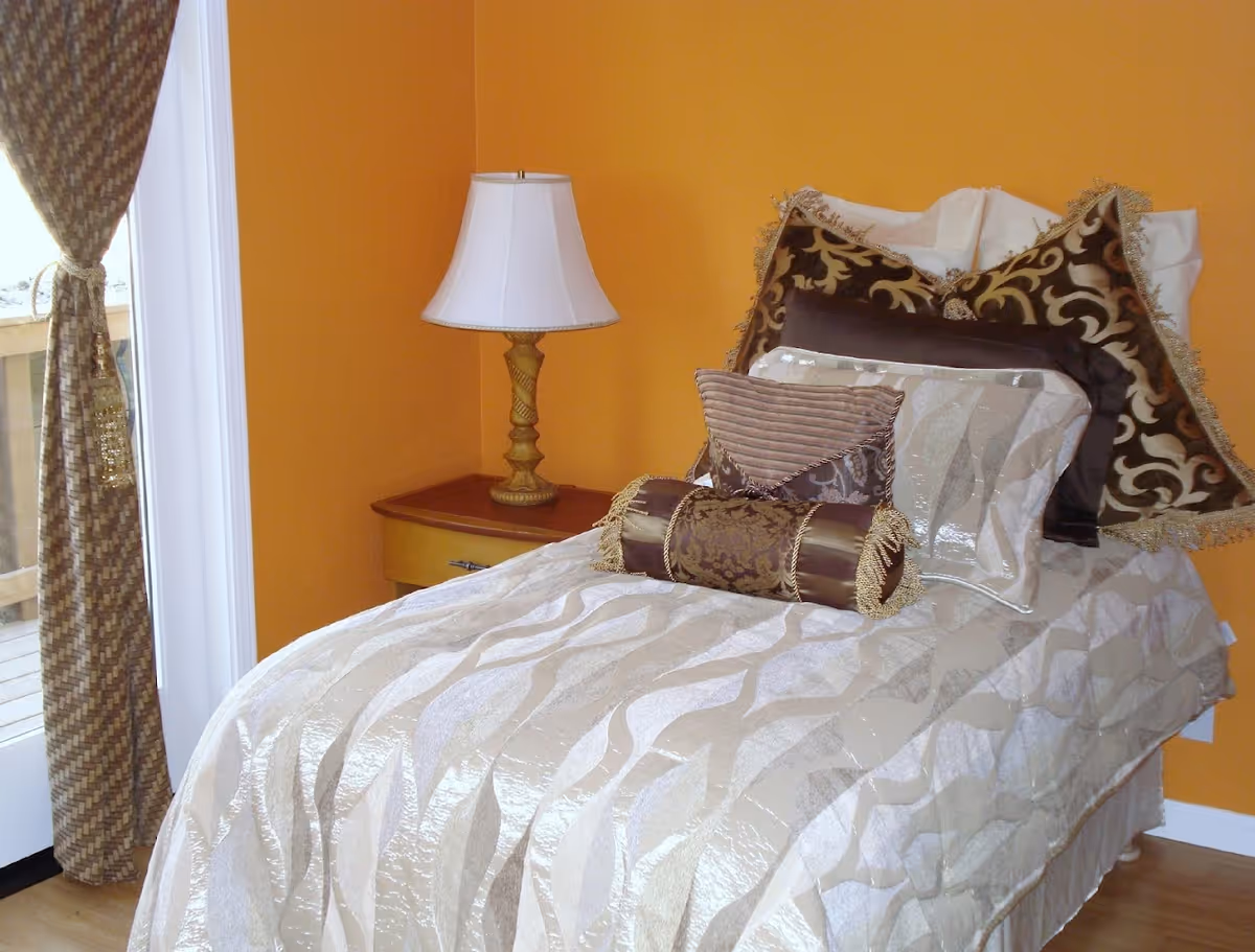 A neatly made single bed with multiple decorative pillows in shades of brown and beige, placed against an orange wall. Next to the bed is a wooden nightstand with a classic table lamp. A window with patterned curtains tied back is visible on the left side, allowing natural light into the room.