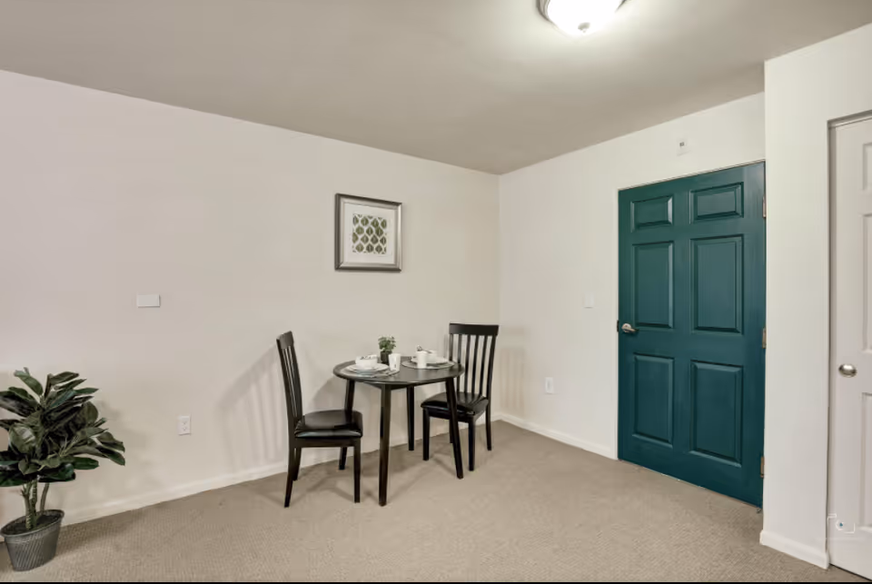 A small dining area with a round black table set for two with white cups and plates. Two black chairs are placed around the table. There is a green potted plant on the floor to the left and a framed artwork on the white wall behind the table. A dark green door and a white door are visible on the right side of the image.