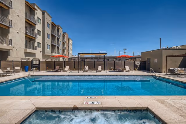 Outdoor swimming pool area at 55 Resort Apartments Downtown Greeley with lounge chairs, umbrellas, and a hot tub in the foreground under a clear blue sky.