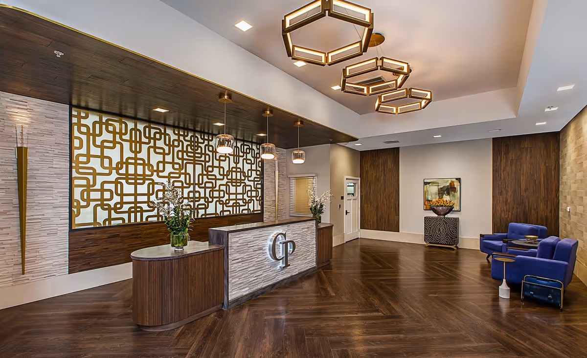 A modern reception area with a wooden floor and a front desk featuring the letters 'CP'. Behind the desk is a decorative wall with a geometric gold pattern and three pendant lights hanging from a dark wooden ceiling panel. To the right, there are two blue armchairs with a small table between them, a decorative cabinet with a bowl of flowers, and a framed abstract painting on the wall. The ceiling has three hexagonal light fixtures.