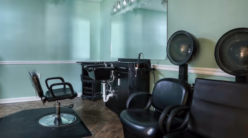 Interior of a salon area with a black salon chair in front of a black sink and counter, two black hair dryer chairs with hooded dryers attached, and light green walls with a large mirror above the sink.