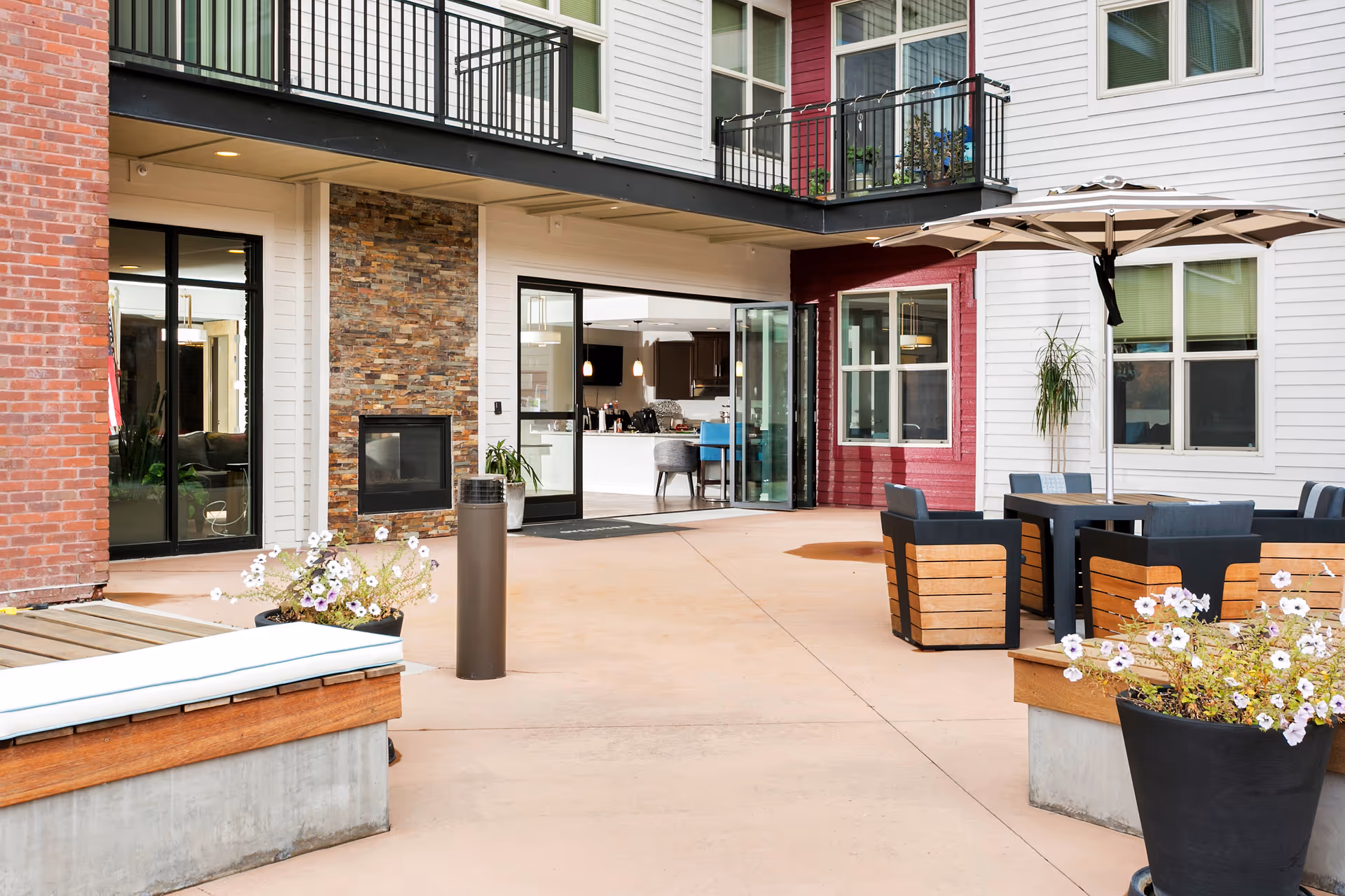 Outdoor patio area at Overture Central Park featuring seating with wooden and black cushioned chairs around a table with a large umbrella. There are flower pots with white flowers, a bench with a cushion, and a building facade with white and red siding, black railings on balconies, and large glass doors leading inside to a kitchen and dining area.