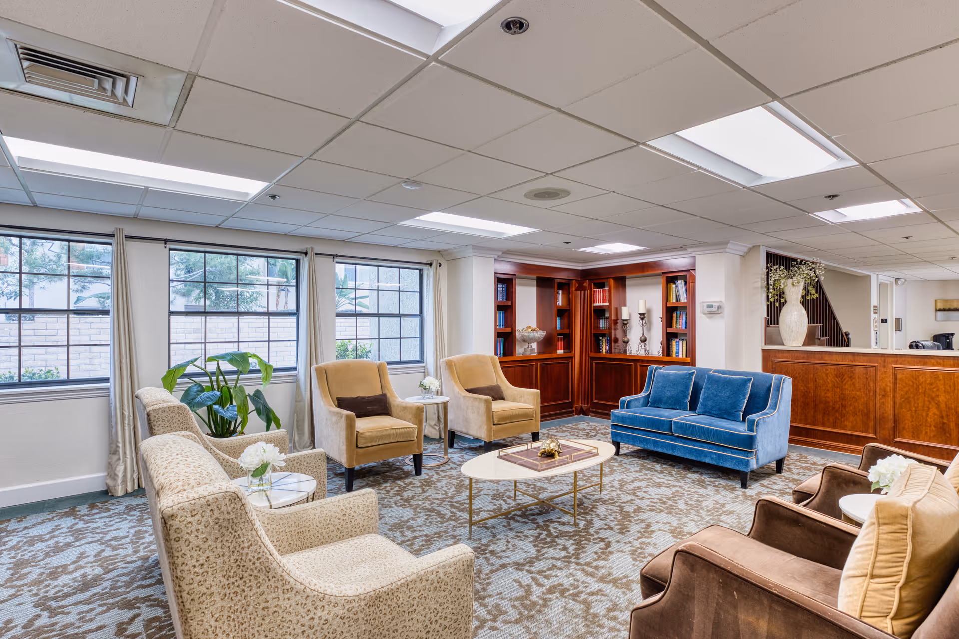 A bright common lounge with assorted armchairs, a blue sofa, coffee table, bookshelves and large windows.