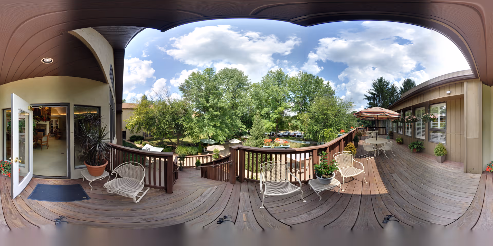 A spacious wooden deck area at Meadowood facility with outdoor seating including metal chairs and tables with umbrellas. The deck overlooks a garden with trees and a parking area in the background. The deck is partially covered with a ceiling and has potted plants along the edges. An open door leads inside to a room with furniture visible.