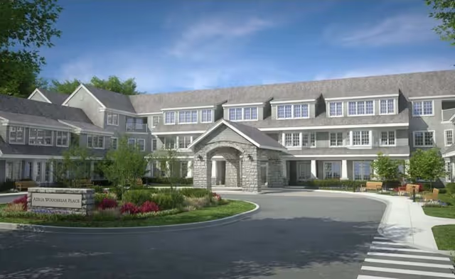 Exterior view of a large senior living facility named Atria Woodbriar Place, featuring a multi-story building with numerous windows, a covered entrance with stone pillars, landscaped greenery, and a circular driveway.