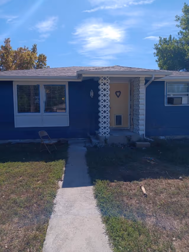 Front exterior of a small blue single-story house with a yellow door and a concrete walkway leading to it.