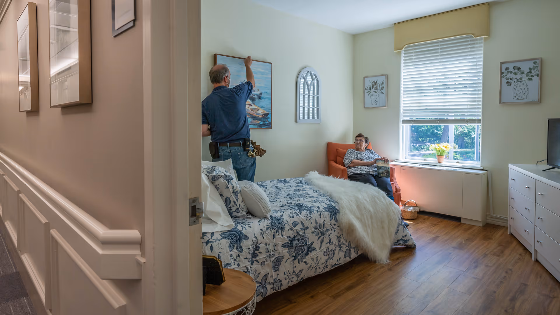 A senior woman sitting in an orange armchair by a window with blinds, holding a book. A man wearing a mask is hanging a painting of boats on the wall above a bed with blue and white floral bedding and a white fluffy throw. The room has wooden flooring, light-colored walls, and framed botanical artwork on the walls.