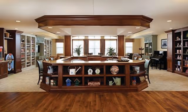 A spacious and well-lit common area in a senior living facility featuring a large wooden central counter with shelves displaying decorative items. Surrounding the counter are several chairs and tables, with bookshelves and a piano visible in the background. The room has large windows with curtains allowing natural light to fill the space.