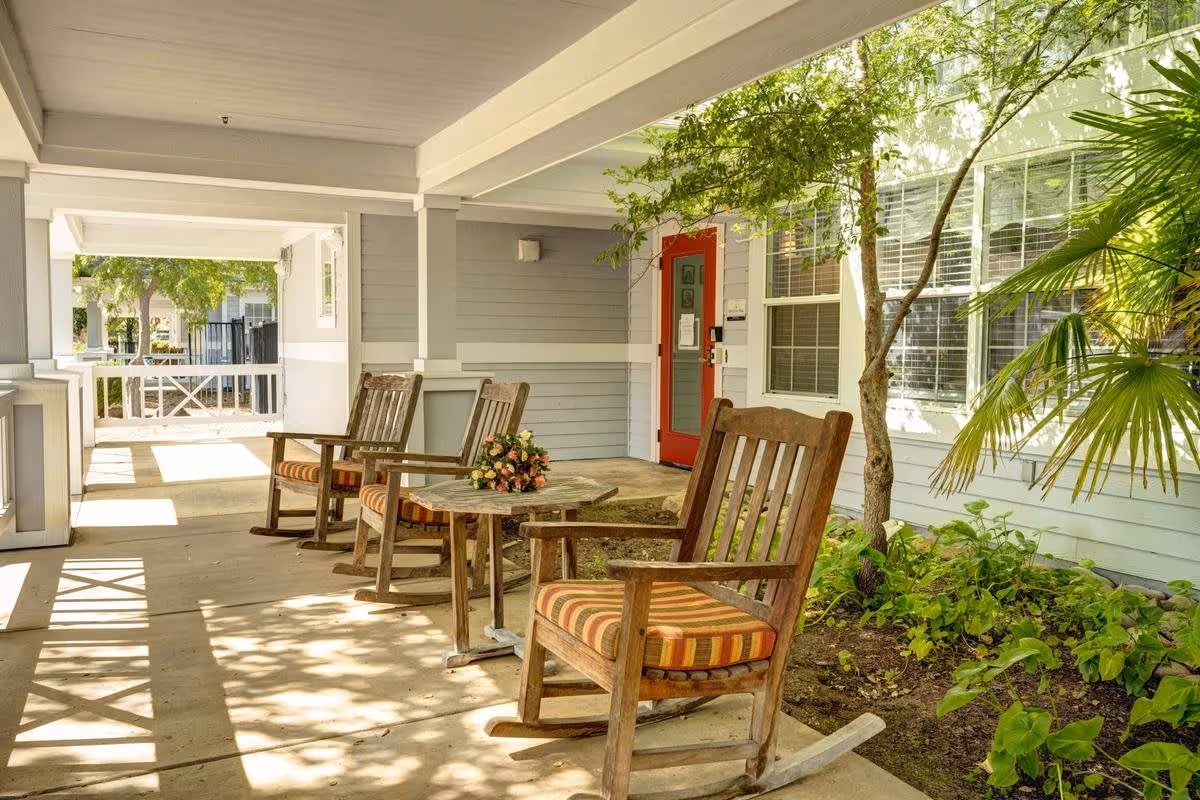Covered outdoor patio area with wooden rocking chairs featuring striped cushions, a small round table with a flower arrangement, and greenery including a tree and plants near the building with a red door and windows.