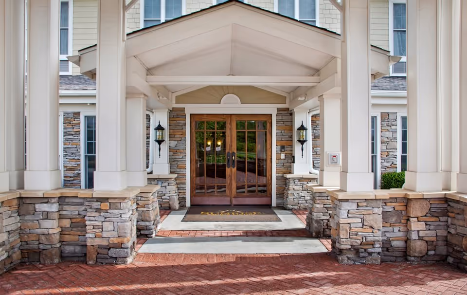 Entrance of a senior living facility with double wooden doors, stone pillars, and a covered porch area. The building exterior features stone and siding with windows visible above the entrance.