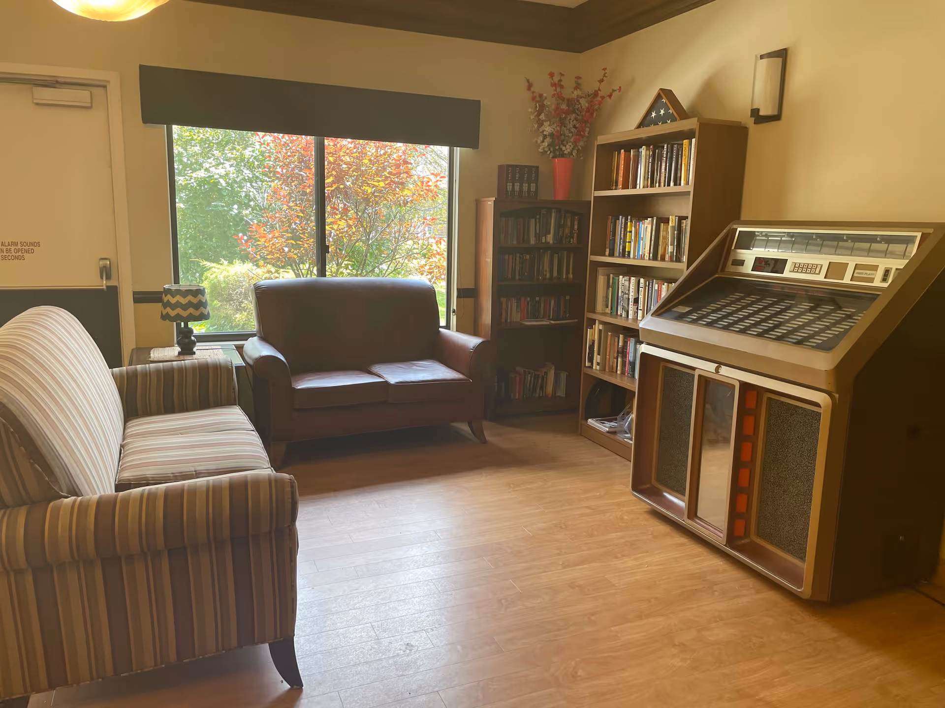 A cozy living room area with a striped sofa and a brown leather loveseat facing a large window showing green and red foliage outside. There is a small table with a lamp between the sofas, a bookshelf filled with books, a smaller bookshelf, a flower vase with red and white flowers, and a vintage jukebox on the right side of the room. The room has wooden flooring and beige walls.