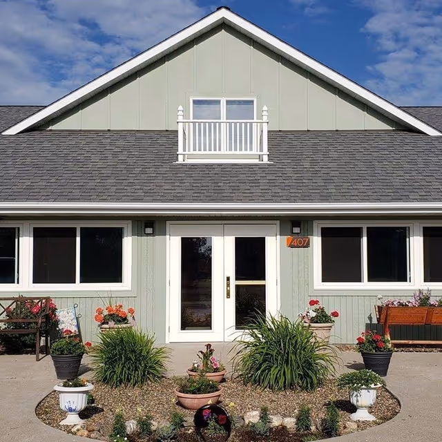 Front exterior view of a light green building with a gray shingled roof, white double doors, two large windows on either side, and a small balcony above the entrance. The entrance is surrounded by a landscaped area with various potted plants and flowers, benches, and a clear blue sky with some clouds in the background.