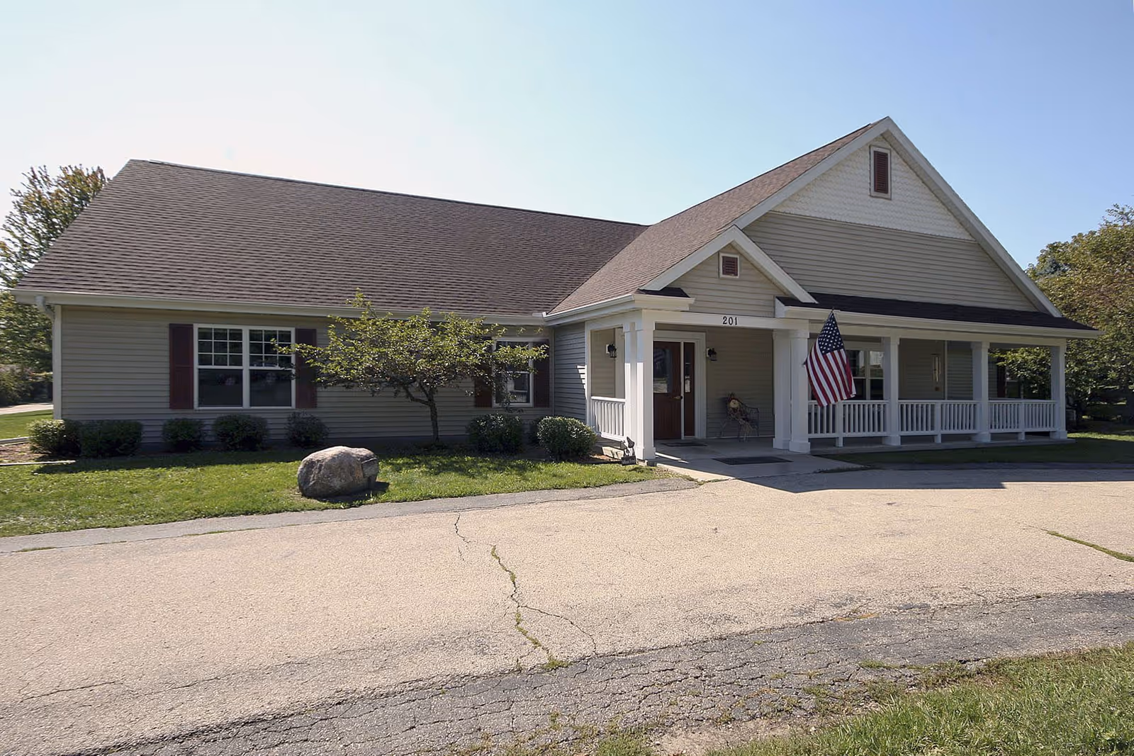 Exterior view of a single-story building with gray siding and a dark shingled roof. The building has a covered porch with white railings and columns, an American flag hanging near the entrance, and the number 201 above the door. There is a small tree and bushes in front of the building, and a paved driveway leading up to it.