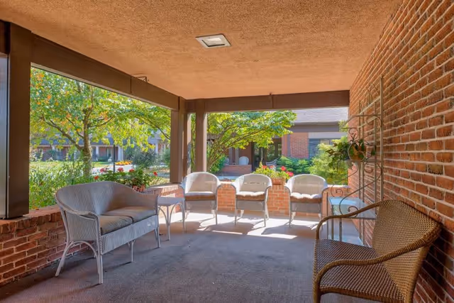 Covered outdoor patio area with wicker seating including a loveseat and four chairs, surrounded by brick walls and columns, overlooking a garden with trees and shrubs in a senior living facility.
