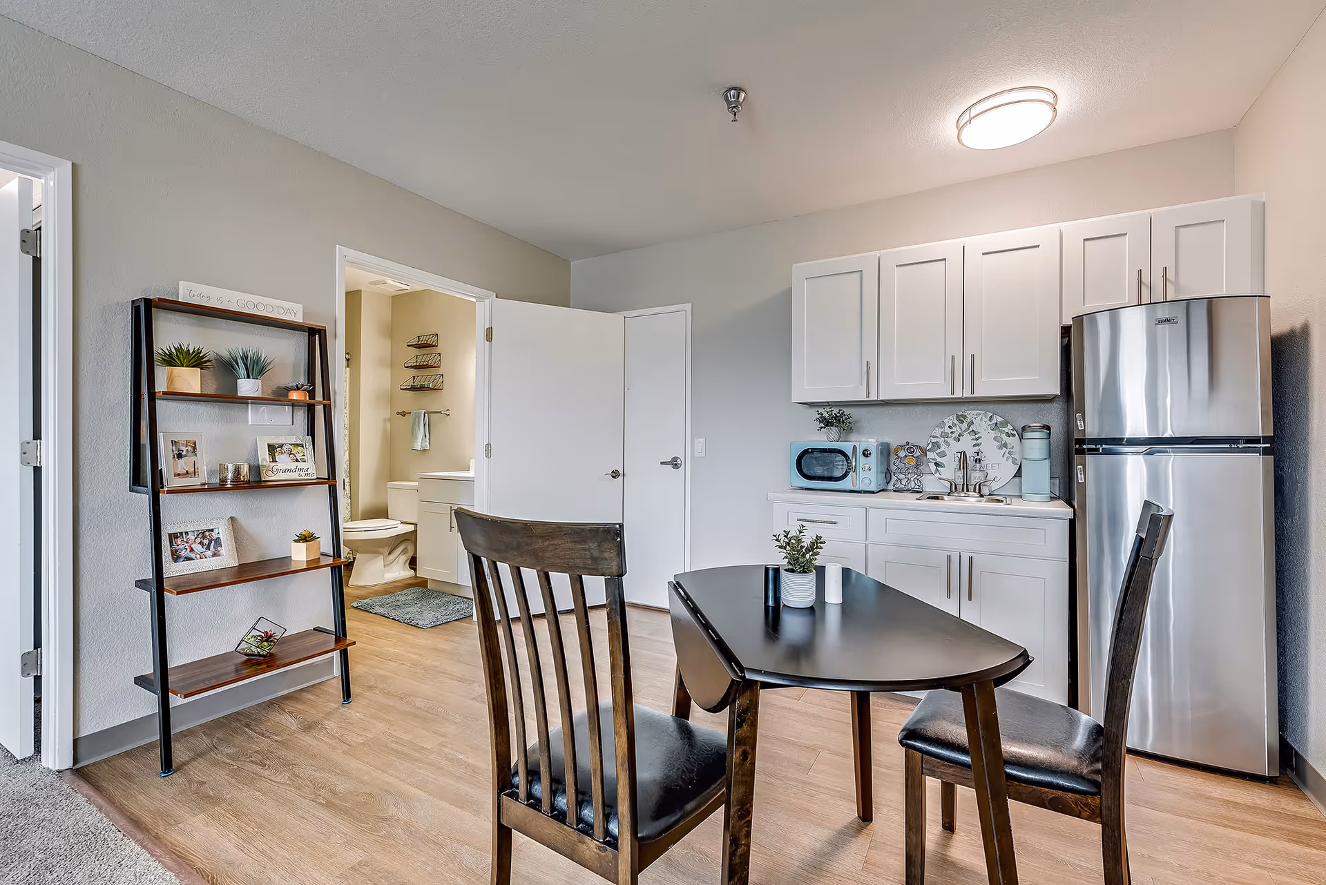Small senior living apartment kitchenette and dining area with a table and chairs, white cabinets, stainless refrigerator, and an open bathroom door.