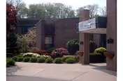 Exterior view of Vernon Rehabilitation and Healthcare Center showing the entrance with a covered walkway supported by brick pillars, surrounded by landscaped bushes and trees.