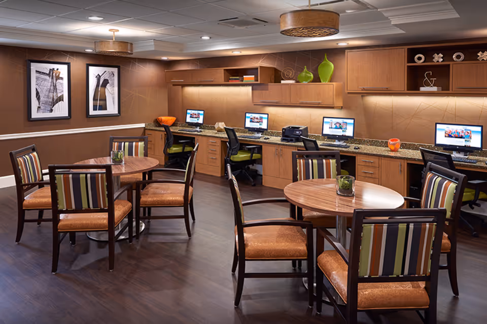 Community seating area with round wooden tables and striped chairs and a row of computer workstations along the back wall.