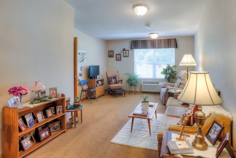 A cozy living room in a senior living facility with beige carpet and walls. The room features a sofa, armchair, coffee table with a plant, side table with a lamp, and a wooden shelf filled with framed photos and a vase of flowers. A window with blinds and a valance lets in natural light, and there are additional chairs and a small TV on a stand in the background.